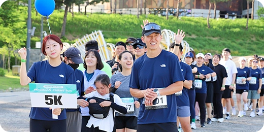 배번호를 단 참가자들이 출발선에 모여 손을 들고 웃고 있습니다. 앞쪽 참가자가 5K 배번호를 들고 있습니다.